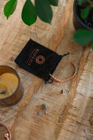 Kopero Bracelet en cuivre Torsadé, sur une table en bois à côté de la pochette en velours, d'une bougie et de bijoux
