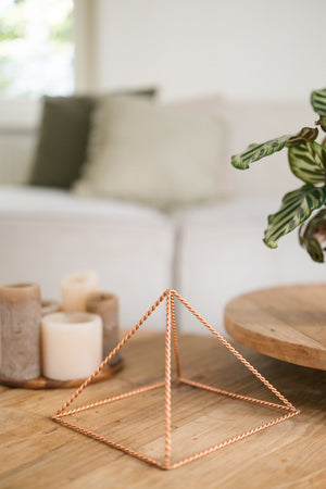 Kopero Pyramide énergisante en cuivre torsadé Grande (23,56 cm), sur une table basse en bois devant un canapé, à côté de bougies