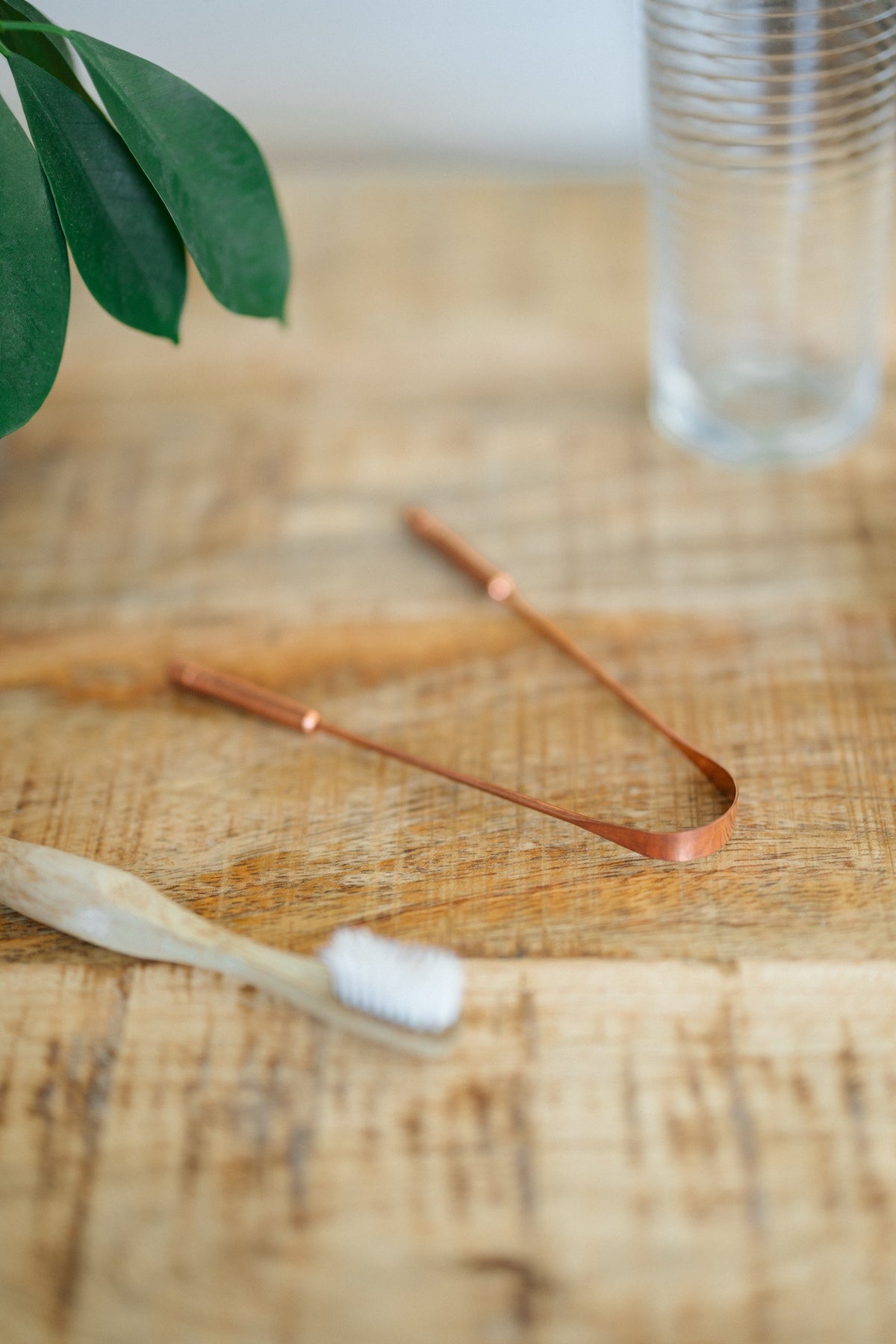 Kopero Gratte-langue en cuivre, sur une table en bois à côté d'une brosse à dents en bambou et d'un verre d'eau