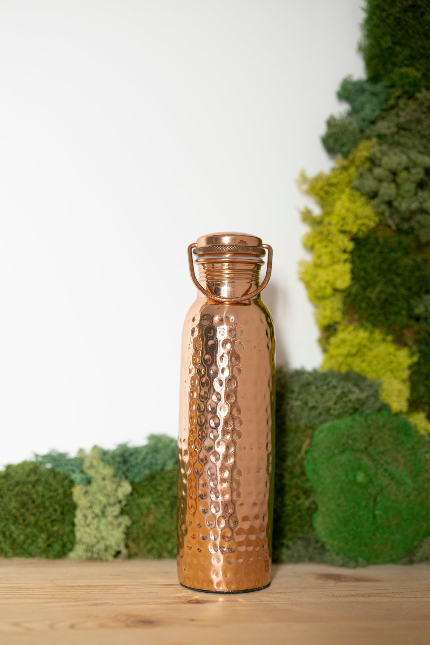 Gourde en cuivre Kopero Martelée, 900 ml, debout sur une table en bois devant un fond de mousse