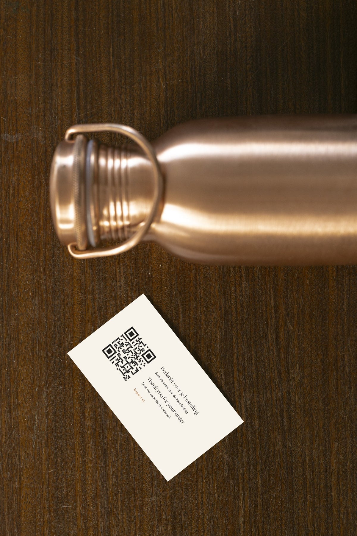 Bouteille d'eau en cuivre Kopero Lisse posée sur du bois, avec le bouchon détaché et une carte de remerciement